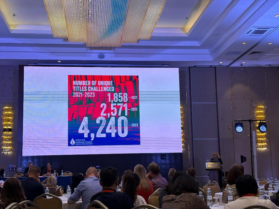 Lidé v přednáškovém sále sledují promítací plátno, vpravo u řečnického pultu stojí žena s brýlemi